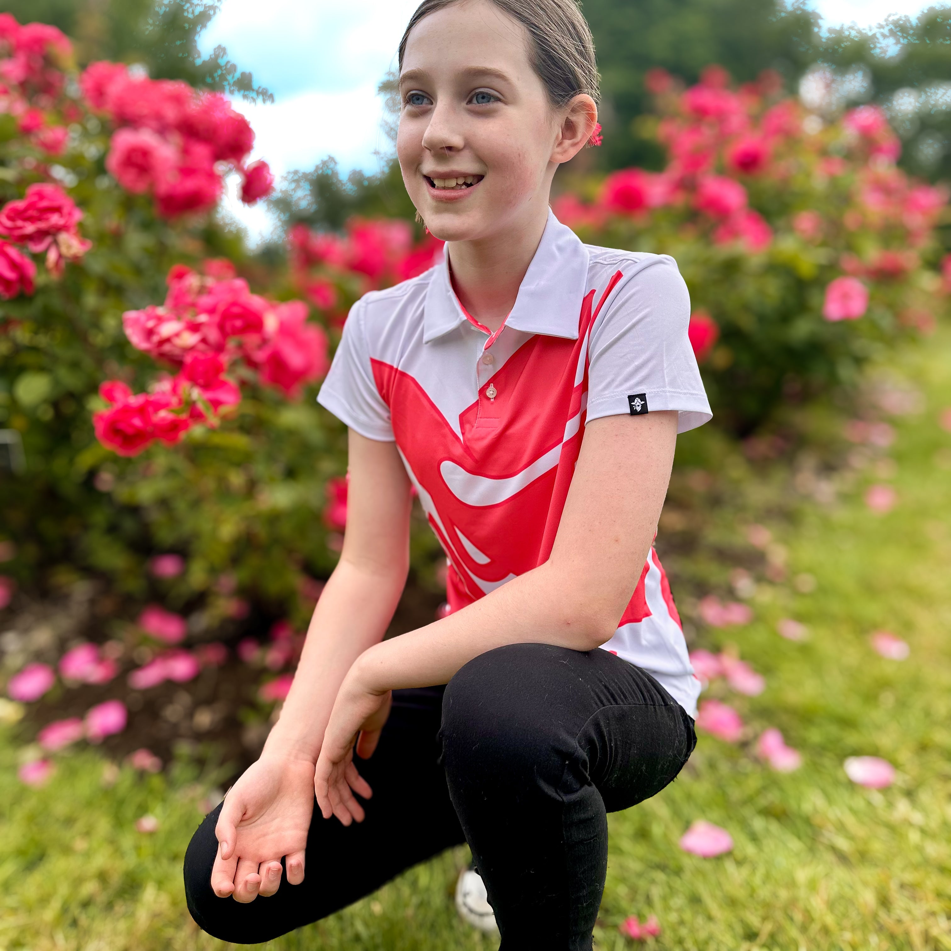 Girl wearing Bandita Blossom Girls Junior Golf Polo in bold pink and white graphic design, posing in a garden with pink flowers. Showcasing moisture-wicking fabric, SPF 50 protection, wrinkle-resistance, and 4-way stretch. Perfect junior golf clothing for girls aged 8-14. Golf Bandit Junior Golf Polos are the best quality in US Kids Golf apparel.