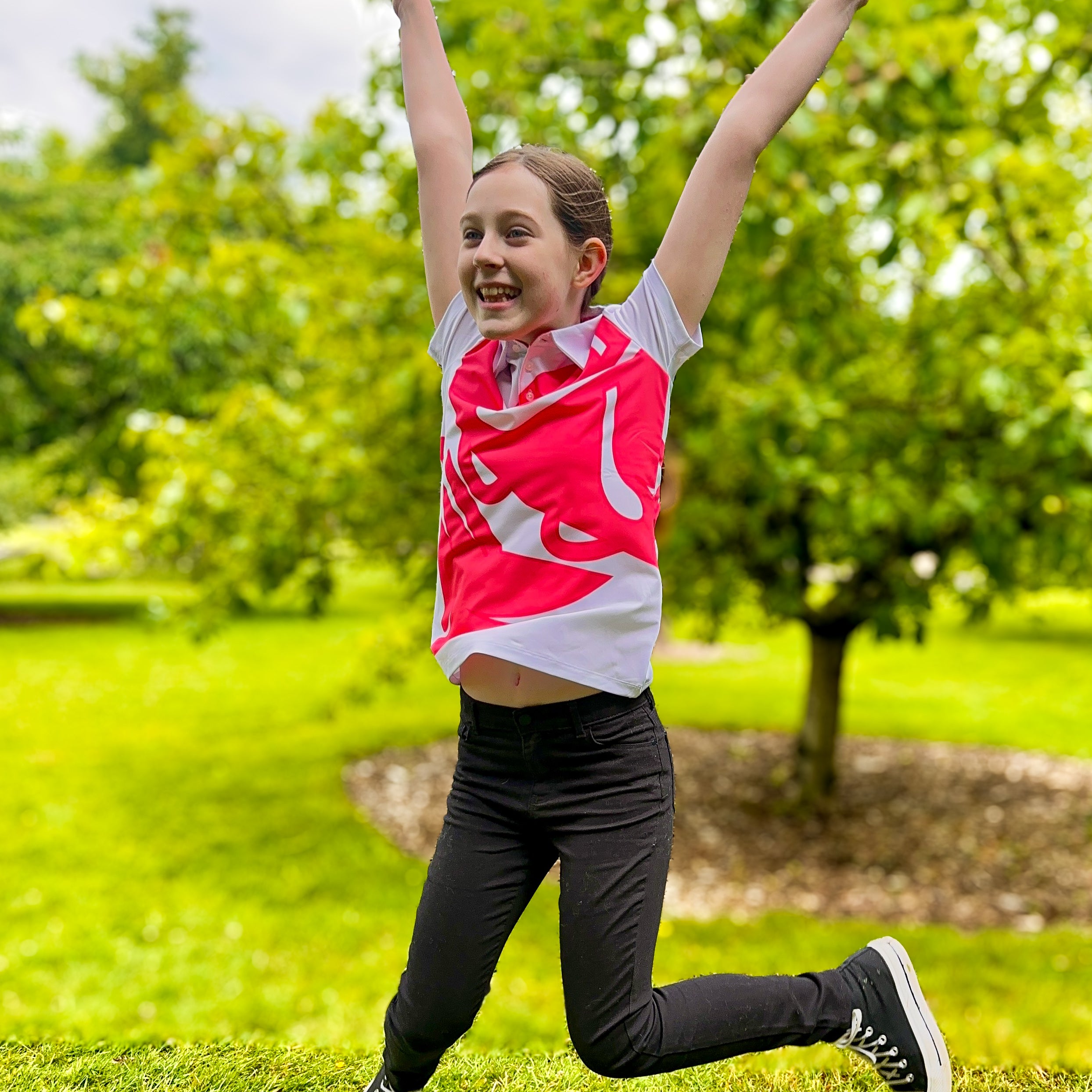 Girl jumping in excitement while wearing Bandita Blossom Girls Junior Golf Polo in bold pink and white graphic design. Showcasing moisture-wicking fabric, SPF 50 protection, wrinkle-resistance, and 4-way stretch. Perfect junior golf clothing for girls aged 8-14. Golf Bandit Junior Golf Polos are the best quality in US Kids Golf apparel.