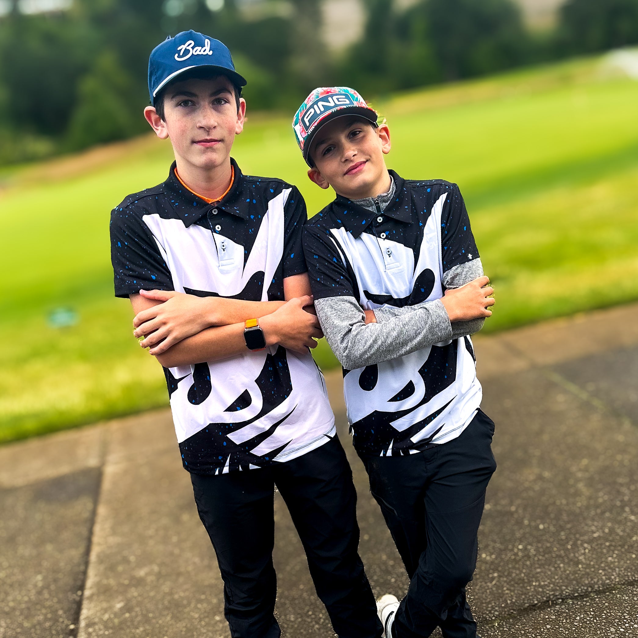 Two boys wearing Bandito Blitz Boys Junior Golf Polo in bold black and white graphic design with blue speckles. Showcasing moisture-wicking fabric, SPF 50 protection, wrinkle-resistance, and 4-way stretch. Perfect junior golf clothing for boys aged 8-14. Golf Bandit Junior Golf Polos are the best quality in US Kids Golf apparel.