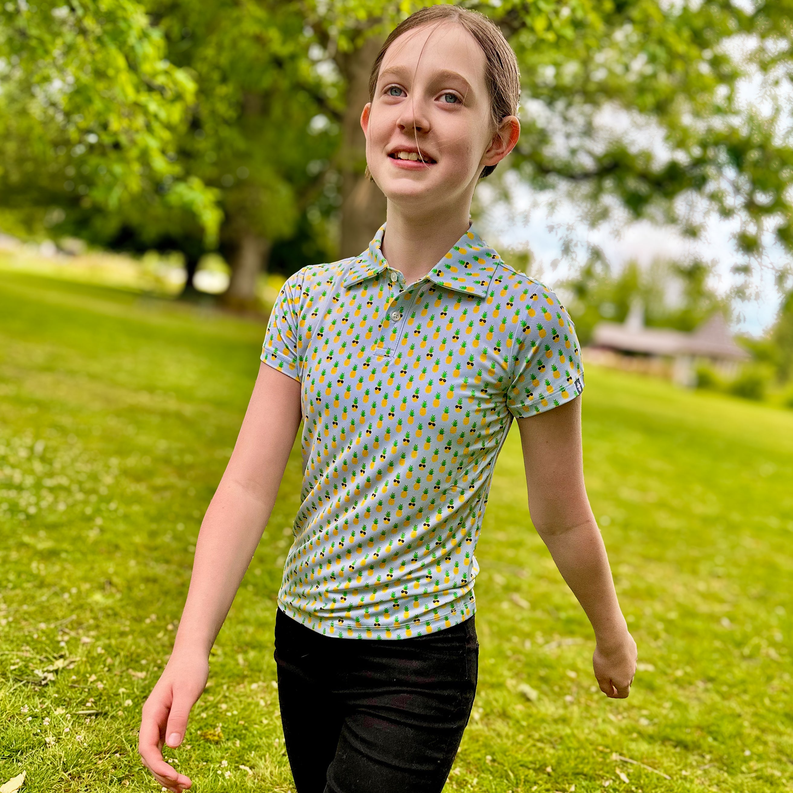 Girl wearing La Piña Girls Junior Golf Polo in light blue with playful pineapple print, showcasing moisture-wicking fabric, SPF 50 protection, wrinkle-resistance, and 4-way stretch. Perfect junior golf clothing for girls aged 8-14. Golf Bandit Junior Golf Polos are the best quality in US Kids Golf apparel.